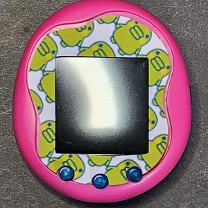 May include: A pink handheld electronic pet toy with a black and white screen. The toy has a white background with a repeating pattern of yellow cartoon ducks. The toy has three blue buttons on the bottom.