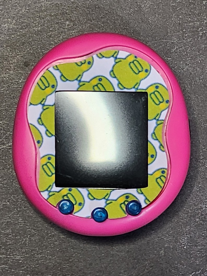 May include: A pink handheld electronic pet toy with a black and white screen. The toy has a white background with a repeating pattern of yellow cartoon ducks. The toy has three blue buttons on the bottom.