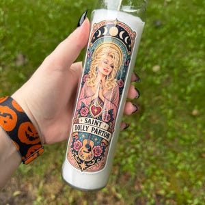 Peut inclure: Une grande bougie de prière cylindrique en verre remplie de cire blanche. L'étiquette présente une illustration de Dolly Parton aux cheveux blonds, vêtue d'une robe rose, les mains jointes, entourée de roses, d'une guitare et de phases lunaires avec le texte SAINT DOLLY PARTON.