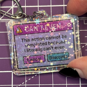 May include: A clear acrylic keychain with a holographic star border. The keychain features a purple and pink graphic with the text "CAN'T EVEN" and a message about an action that cannot be completed. It includes "Cancel" and "Dissociate" buttons.