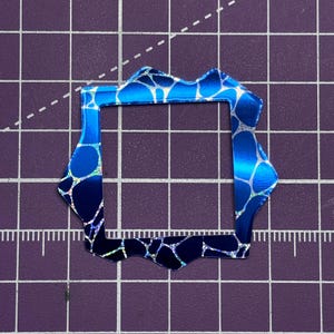May include: A blue and silver decorative frame with a unique, irregular edge. The frame has a square interior and a textured, iridescent pattern. The color transitions from a lighter blue to a darker blue. The frame is on a purple grid.