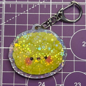 May include: A clear, iridescent keychain featuring a cartoon cantaloupe design. The cantaloupe is light green with a smiling face and red cheeks. The keychain has a silver clasp and chain.