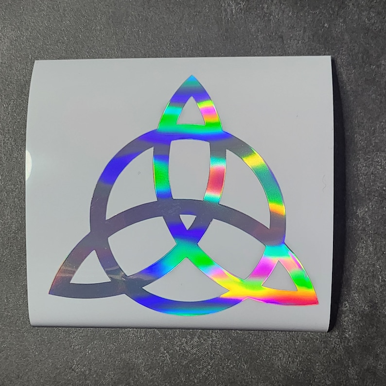 K&ouml;nnte beinhalten: Ein silberner holografischer Vinyl-Aufkleber mit einem Triquetra-Design. Die Triquetra ist ein Symbol f&uuml;r drei miteinander verbundene Kreise, die oft mit Spiritualit&auml;t und der Trinit&auml;t in Verbindung gebracht werden.