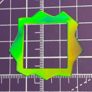 May include: A neon green and yellow iridescent square frame with an irregular, jagged outer edge. The frame has a square cut-out in the center. The frame is on a purple grid background.
