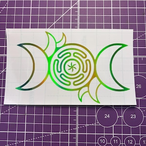 May include: A holographic sticker featuring a triple moon symbol with a central spiral design and a star in the middle. The sticker has a gradient of green and gold colors, with a white background.