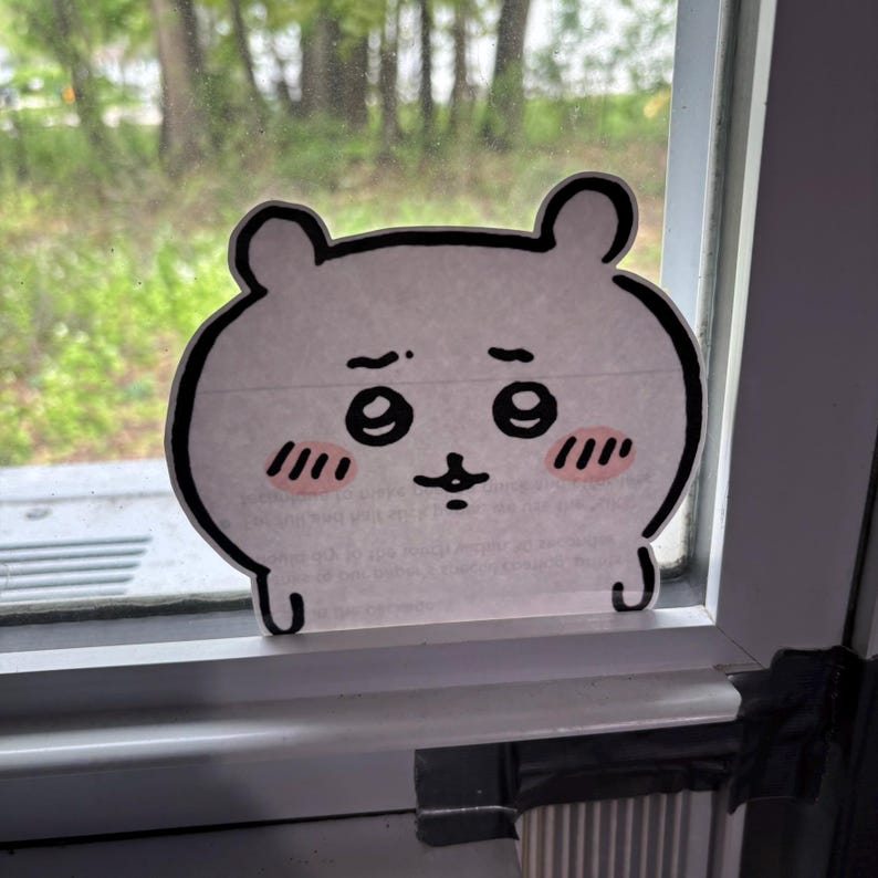K&ouml;nnte beinhalten: Ein wei&szlig;es Comic-Tier mit schwarzen Augen und einem rosa Rouge auf den Wangen. Das Tier ist auf ein Blatt Papier gezeichnet und an einem Fenster befestigt.