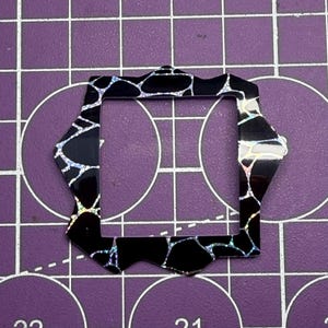 May include: A black, geometric craft element with an iridescent pattern. The shape is a modified octagon with a square opening. The background is a purple cutting mat with grid lines and circular markings.
