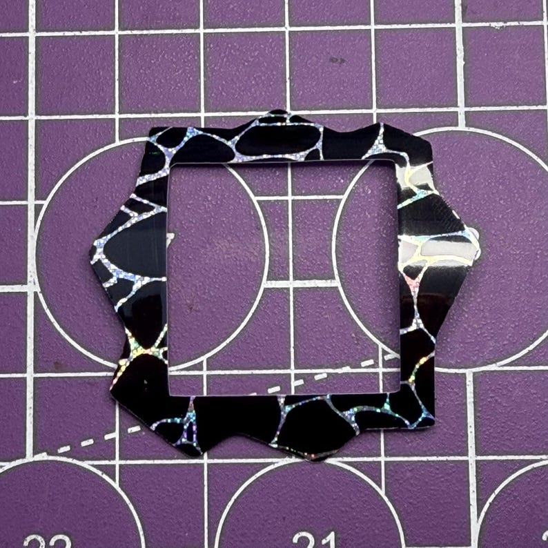 May include: A black, geometric craft element with an iridescent pattern. The shape is a modified octagon with a square opening. The background is a purple cutting mat with grid lines and circular markings.