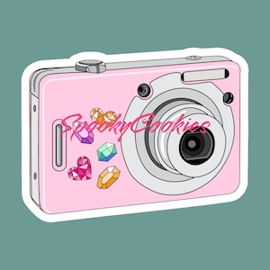 May include: A pink digital camera with a silver lens and a white border. The camera has several colorful gems and rhinestones on the front.