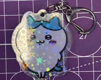 May include: A clear, holographic cat-shaped keychain with a silver clasp and key ring. The cat has blue accents on its head and paws, with a smiling face and star-shaped details. The background is a purple grid.