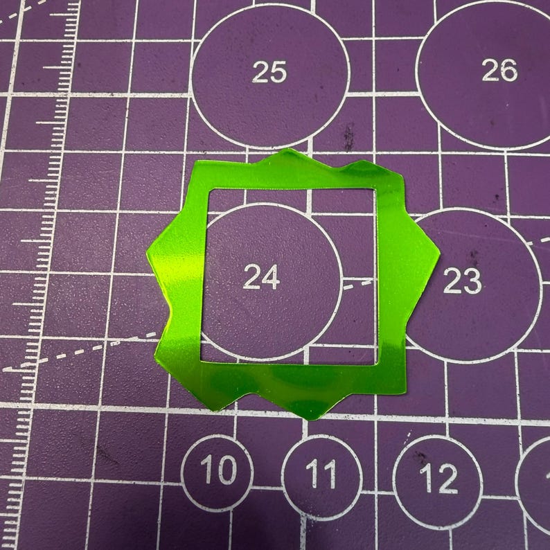 May include: A bright green, reflective, star-shaped cutout with a square center hole, positioned on a purple cutting mat with white grid lines and circular markings, including the number 24 visible through the center of the cutout.