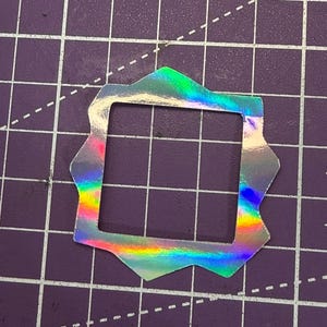 May include: A holographic sticker with a square center and an ornate, multi-pointed border. The sticker reflects a rainbow of colors, including green, blue, yellow, and pink. It is placed on a purple cutting mat with grid lines.