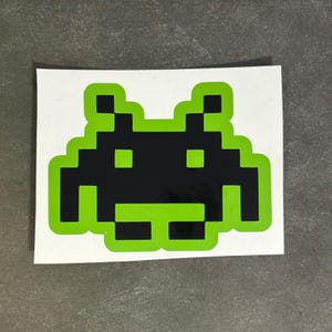 May include: A green and black pixelated alien sticker. The alien has a rounded body with two antennas and two eyes.