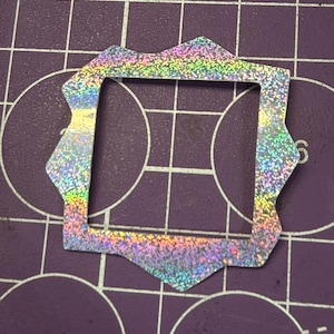 May include: A holographic, iridescent square-shaped sticker with a unique, scalloped edge. The sticker reflects a rainbow of colors, including pink, yellow, green, and blue. The sticker is placed on a purple cutting mat with white grid lines and circular markings.