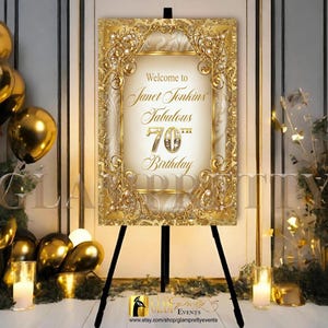 May include: A rectangular welcome sign on a black easel featuring a gold ornate frame with pearl accents. The center text reads Welcome to Janet Tonkins Fabulous 70th Birthday in elegant gold script and serif fonts against a cream background.