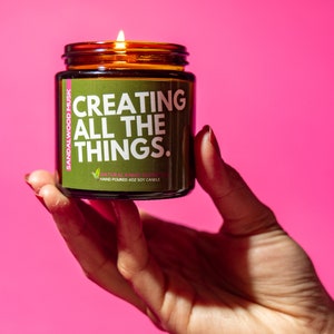 May include: A brown glass candle jar with a green label that reads "Creating All The Things."  The candle is lit and the label also says "Sandalwood Musk" and "Natural Plant-Based Essential Oils" and "Hand Poured 4oz Soy Candle".