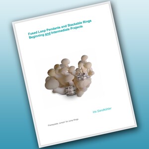 May include: A white paper with the title "Fused Loop Pendants and Stackable Rings Beginning and Intermediate Projects" and the text "Prerequsite: Jumpin' for Jump Rings" and the name "Iris Sandkühler". The image also shows a cluster of white mushrooms with silver rings on top.