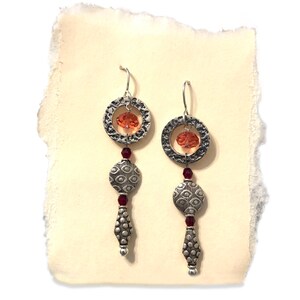 May include: Silver dangle earrings with a textured circular design, featuring a small orange crystal bead and a larger round bead with a patterned design. The earrings are accented with small red crystal beads.