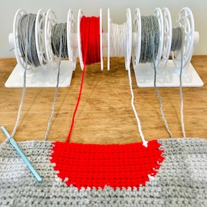 May include: A crochet project in progress, with a colour scheme of grey and red. White yarn spools are arranged in the centre, with grey yarn spools on either side. A crochet hook lies on the grey crocheted fabric, next to a red section.