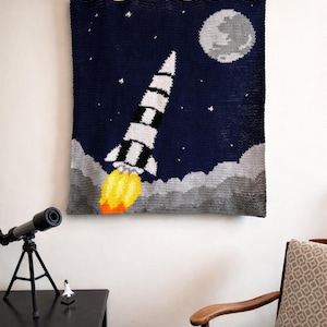 May include: A navy blue, gray, and white crocheted wall hanging depicting a rocket ship blasting off into space. The rocket has black and white stripes, with yellow and orange flames. A gray moon and stars are in the background.