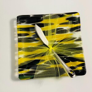 May include: A square glass dish with a yellow and black striped design, a silver knife, and a yellow mesh ribbon. The dish is approximately 5 inches square. The knife has a decorative end. The overall theme is a decorative serving piece.