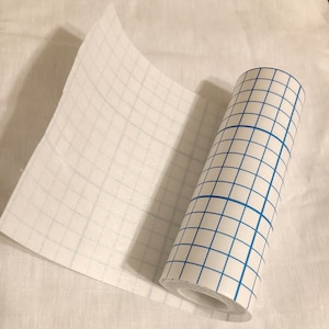 May include: A roll of white medical tape with a blue grid pattern. The tape is partially unrolled and the roll is lying on a white surface.