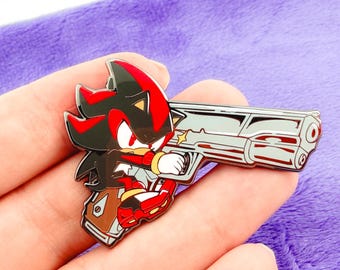 May include: A colorful enamel pin featuring a cartoon character holding a silver gun. The character has black and red spiky hair, red shoes, and a white glove. The pin is held against a purple background.
