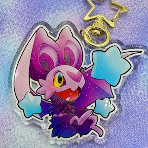 Puede incluir: Un llavero acrílico transparente con una criatura de dibujos animados con forma de murciélago con detalles en morado, rosa y azul. El llavero tiene un cierre dorado en forma de estrella y varios adornos azules en forma de estrella. El fondo es de tela morada clara.