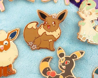 May include: A collection of metal enamel pins featuring various stylized creatures with floral accents. The pins include a brown Eevee with daisies, a black and yellow Umbreon with red roses, a cream-colored Leafeon, and other colorful character designs.
