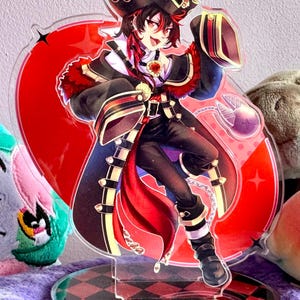 Puede incluir: Un colorido soporte acrílico con un personaje de dibujos animados con un atuendo de temática pirata. El personaje lleva un sombrero negro con detalles dorados, una capa roja y pantalones negros. El soporte tiene una base a cuadros roja y negra.