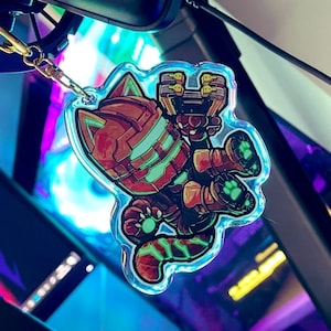Könnte beinhalten: Ein klarer Acryl-Schlüsselanhänger mit einer Cartoon-Katze in einem Roboteranzug. Die Katze ist hauptsächlich braun und grün, mit einem goldfarbenen Verschluss. Der Schlüsselanhänger hat einen holografischen Rand.