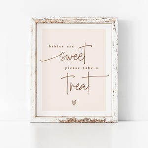 Op de afbeelding: Een wit ingelijst bord met een lichtroze achtergrond en bruine tekst die "baby's zijn zoet, neem een traktatie" zegt met een klein hartje onderaan.