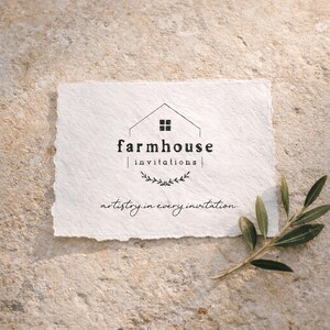 May include: A rectangular piece of textured white paper with the words "farmhouse invitations" in black. Below the text is a small leaf design. The phrase "artistry in every invitation" is written in a script font. A sprig of green leaves is on the surface.