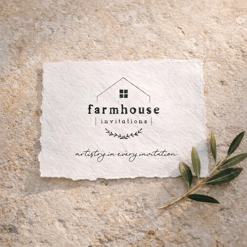 May include: A rectangular piece of textured white paper with the words "farmhouse invitations" in black. Below the text is a small leaf design. The phrase "artistry in every invitation" is written in a script font. A sprig of green leaves is on the surface.