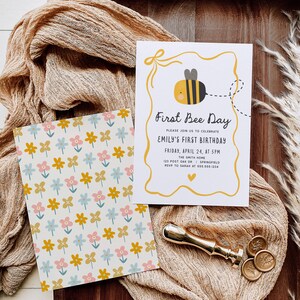 May include: Two cards with a bee theme. One card has a floral pattern in yellow, pink, and blue. The other card says "First Bee Day" with a bee graphic. A gold wax seal and stamp are also present.