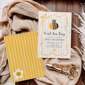 May include: Two-sided invitation with a bee theme. The front features a cartoon bee and the text "First Bee Day" with event details. The back has yellow stripes and a white daisy. A gold wax seal and a beige cloth are in the background.