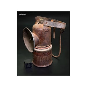May include: An antique, rusty metal miner's lamp with a large reflector and a handle. The lamp has a cylindrical body with a circular base and a handle. The text "6-WGY" is visible on the top left.
