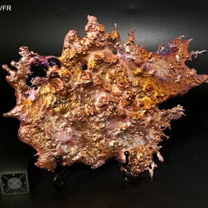 May include: A large, irregularly shaped piece of copper metal with a textured surface. The metal has a variety of colors, including gold, brown, and purple. The piece is displayed on a black metal stand.