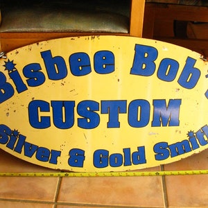 Può includere: Un cartello ovale giallo con scritta blu che recita "Bisbee Bob's Custom Silver & Gold Smith".