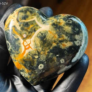 May include: A heart-shaped polished stone with a unique pattern of black, green, white, and orange colors. The stone has a smooth, polished surface.