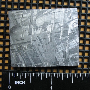 May include: A close-up of a piece of meteorite with a Widmanstätten pattern. The meteorite is a silvery gray color and has a rough texture. The meteorite is about 1 inch wide.