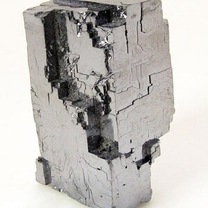 Pu&ograve; includere: Un grande cristallo cubico metallico grigio con una superficie sfaccettata a gradini. Il cristallo ha una forma geometrica e a blocchi.