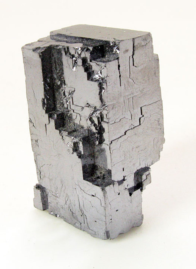 Pu&ograve; includere: Un grande cristallo cubico metallico grigio con una superficie sfaccettata a gradini. Il cristallo ha una forma geometrica e a blocchi.