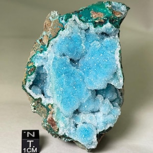 May include: A vibrant, natural mineral specimen featuring a striking contrast of colors. The core showcases a brilliant, crystalline, light blue hue, surrounded by a dark teal and brown matrix. The text "5-G66W" and "1CM" is visible.