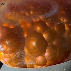 May include: Close-up of a polished, translucent gemstone with a warm, amber hue. The surface features a textured, bubbly pattern with areas of bright orange and yellow, creating a glowing effect. The text "6-EPZZ" is visible in the upper left corner.