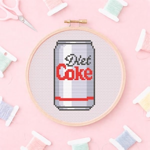 Può includere: Un disegno a punto croce di una lattina di Diet Coke in un telaio da ricamo in legno. La lattina è argentata con testo rosso e una striscia rossa. Forbici rosa e rocchetti di filo di vari colori circondano il telaio su uno sfondo rosa.