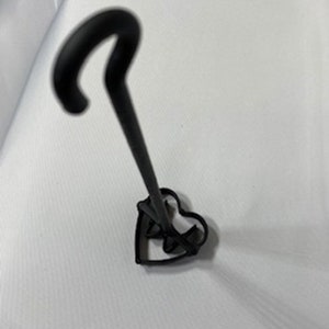 Puede incluir: Cortador de galletas de metal negro en forma de corazón con un mango largo. El mango tiene un gancho en la parte superior.