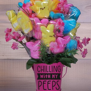 May include: A pink bucket with the text "CHILLING WITH MY PEEPS" in black letters. The bucket is filled with a bouquet of yellow, pink, and blue marshmallow Peeps, surrounded by pink flowers.