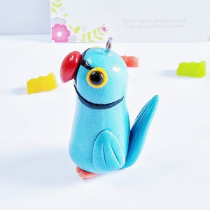 May include: A handcrafted, light blue parrot figurine with a red beak and yellow eyes. The parrot has a black band around its neck and a small metal loop at the top. The background includes colorful candies and a business card with the text "MhavelCraft".