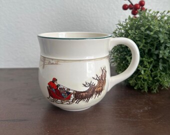 May include: Cream-colored ceramic mug with a green rim and handle. The mug features a festive illustration of Santa Claus in a red sleigh pulled by reindeer. A brown horizontal band separates the top and bottom of the mug.